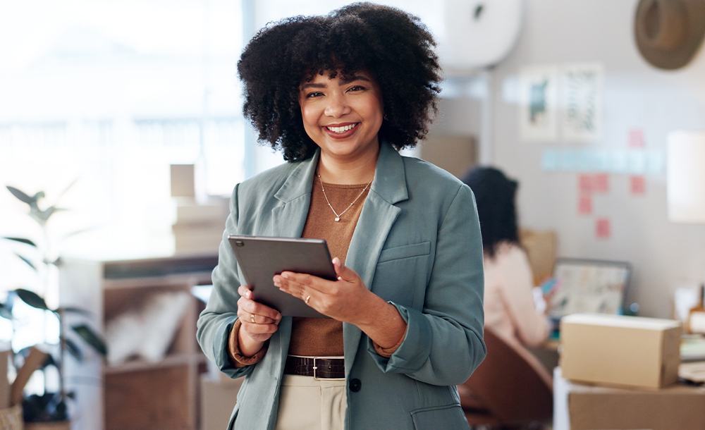 Mulher negra, em ambiente empresarial, sorrindo e segurando um tablet nas mãos.