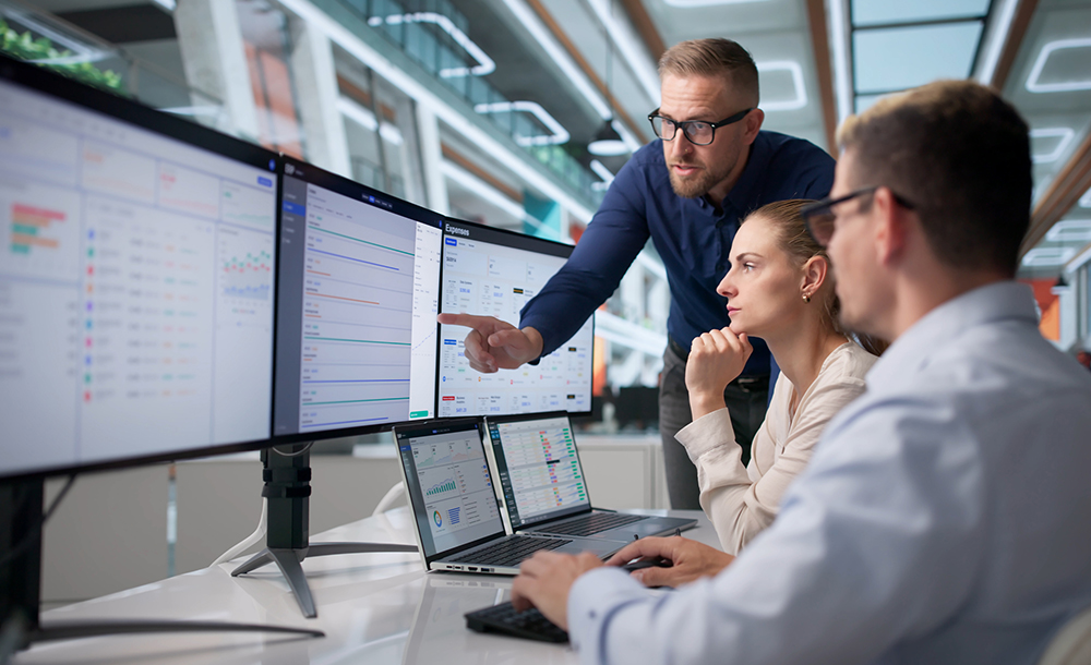 Pessoas em frente a tela de computares, analisando dados, em ambiente empresarial